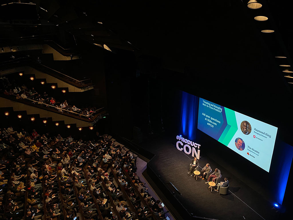 Top view of a convention center with speakers on stage, a PowerPoint presentation on screen, and the audience.